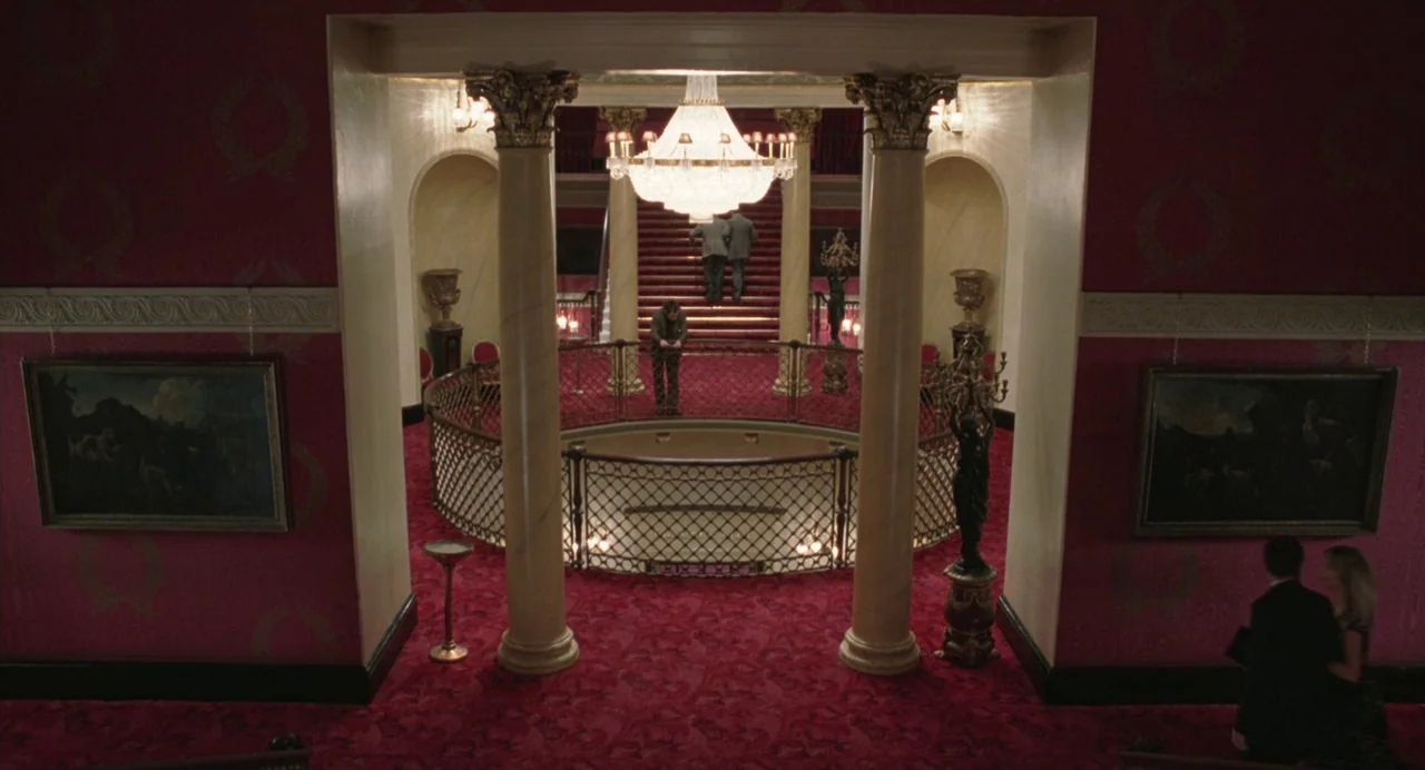 Closer (2004) movie still: wide — A grand interior space features ornate red walls, gold-trimmed columns, and a large chandelier. A man lingers by a…