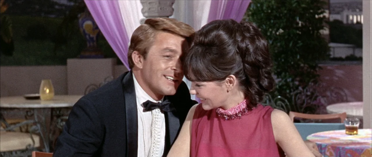 Clambake (1967) movie still: medium — An outdoor restaurant terrace features draped purple fabric and round tables at dusk. A man in a tuxedo leans…
