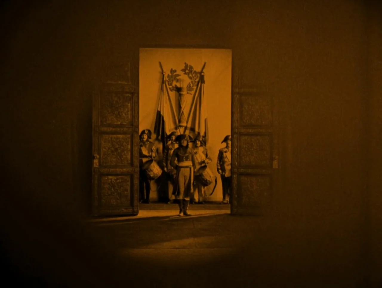 Napoleon (1927) movie still: fullbody — A dark, grand interior space features a central open doorway with a prominent emblem above. A soldier in…