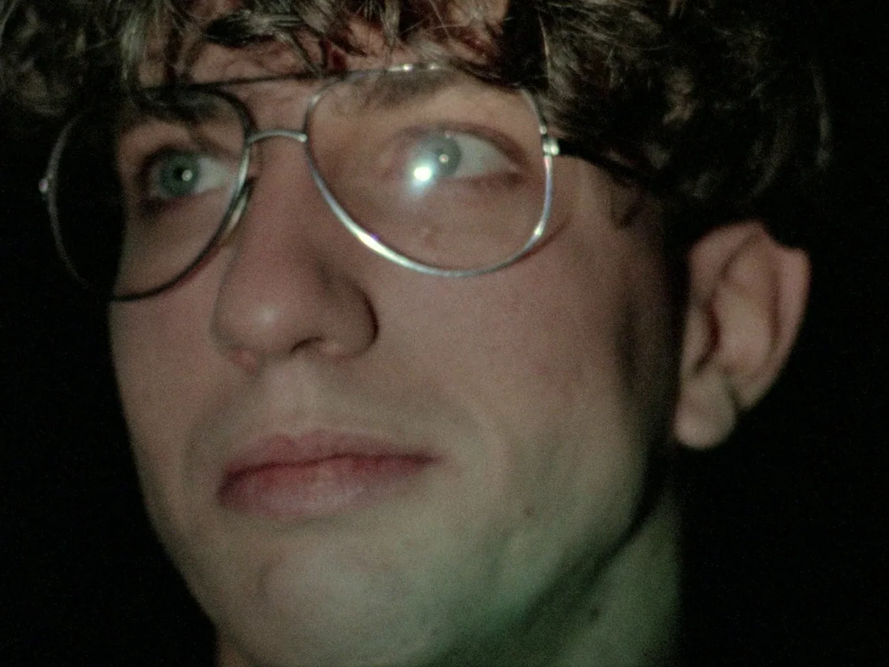 Decoder (1984) movie still: closeup — A close-up shot of a young man with curly hair in a dark environment. The man wears large thin-rimmed glasses…