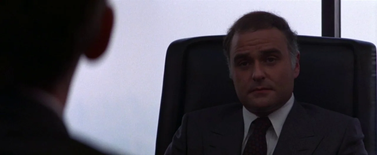 Klute (1971) movie still: medium — A high-rise office interior featuring a large window with bright white daylight background. A man in a suit sits…