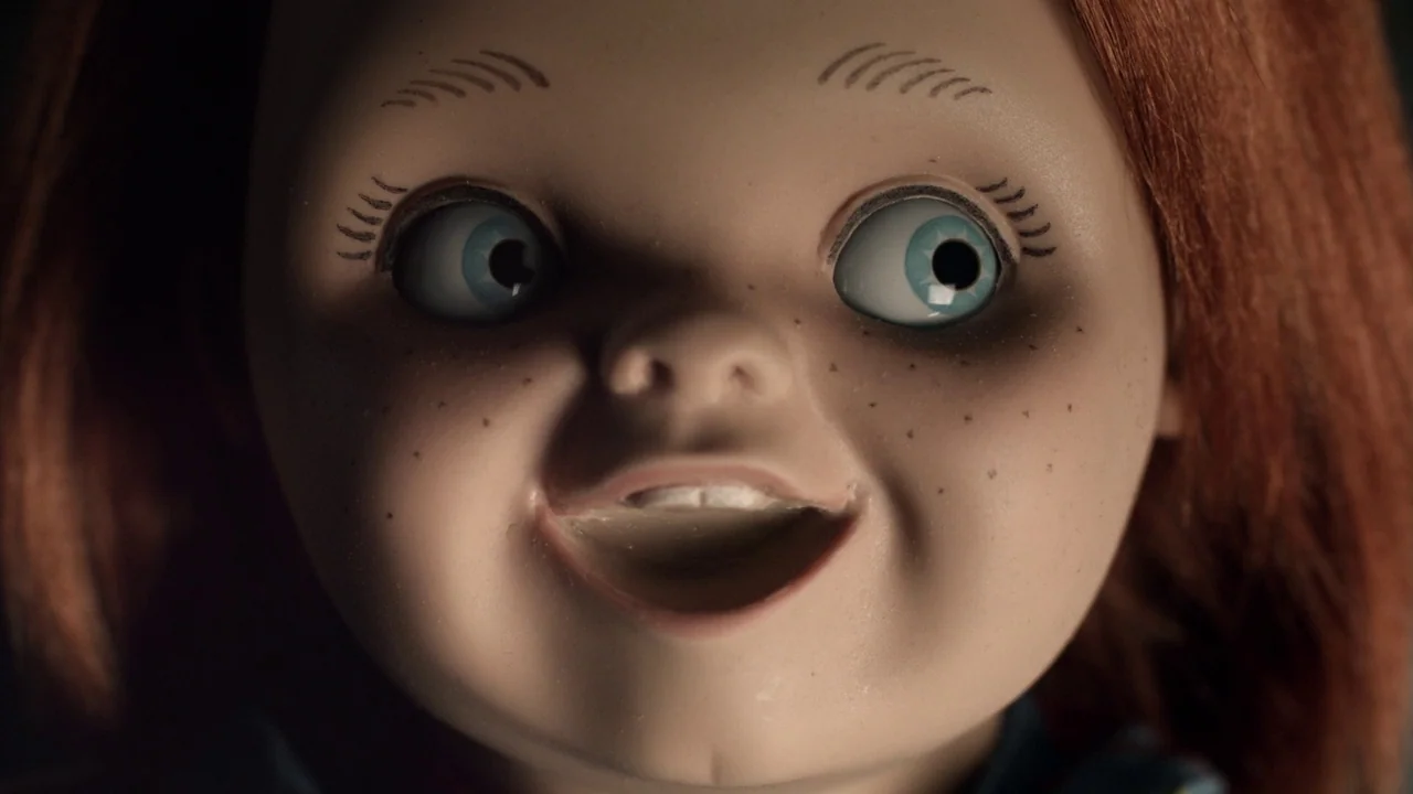 Curse of Chucky (2013) movie still: closeup — A close-up of a doll face with blue eyes and orange hair. The doll stares directly forward against a…