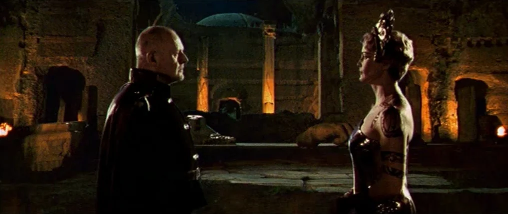 Titus (1999) movie still: medium — An interior space featuring rough stone walls and illuminated ruins in the background. A man in a dark uniform…