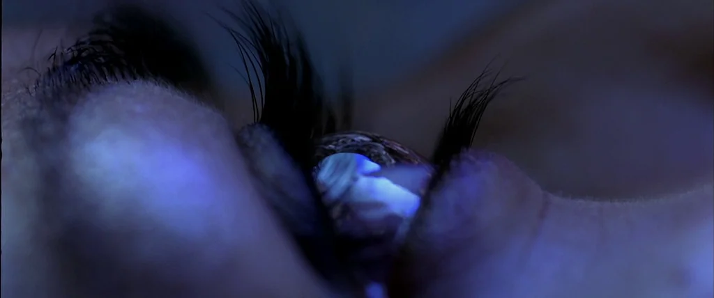 The Cell (2000) movie still: closeup — An extreme close-up of a human eye reflecting a bright blue light source. The eye is partially open with dark…
