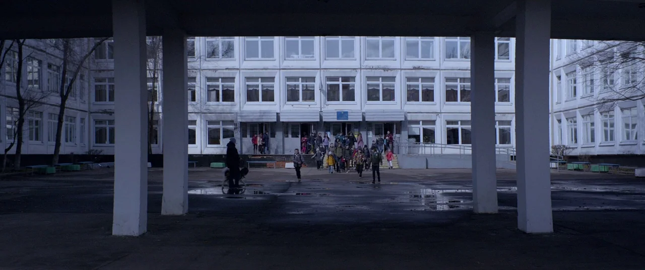 Loveless (2017) movie still: wide — A large concrete school building with many windows sits behind a paved school courtyard. A group of children in…