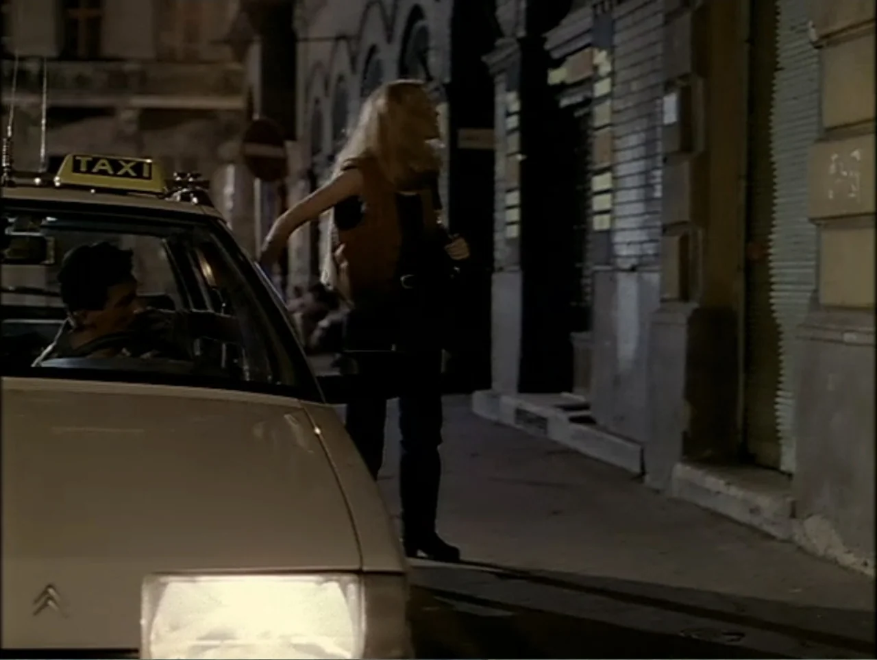 A Change of Place (1994) movie still: medium — A quiet urban street at night features a white taxi parked near brick buildings. A blonde woman…