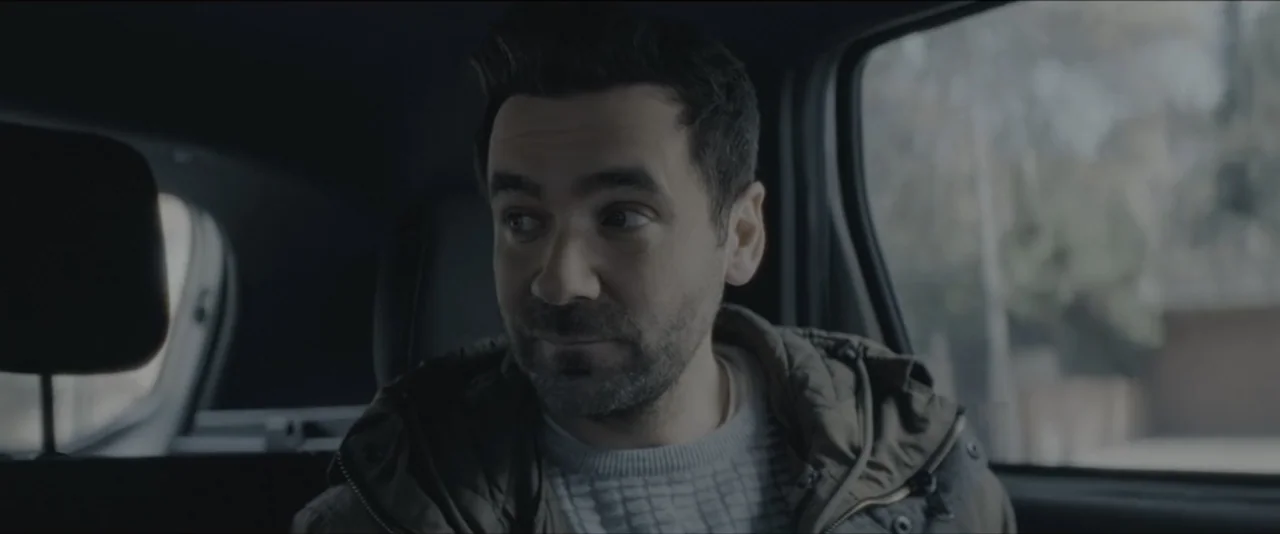 Quicksand (2023) movie still: medium — A man sits in the back of a vehicle during the daytime. He is looking away from the camera toward the window.
