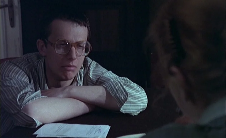 Blind Chance (1987) movie still: medium — An interior space features a dark surface where a person sits behind papers. A man in a striped shirt…