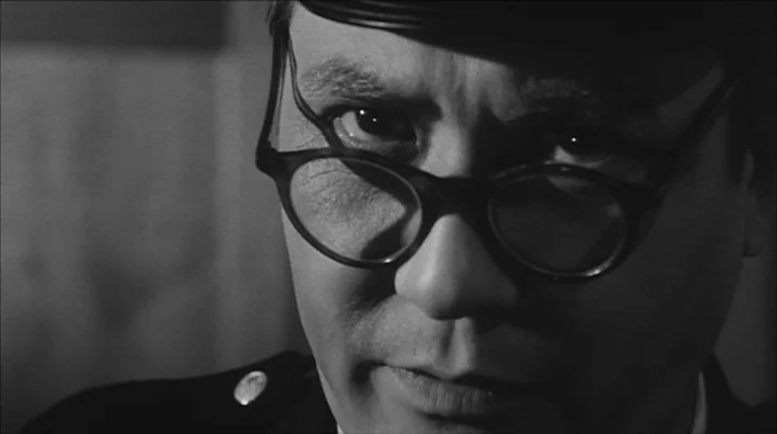 Death By Hanging (1968) movie still: closeup — A man in uniform is visible in close-up against a dark, featureless background. He looks directly at…