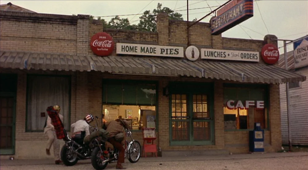 Easy Rider (1969) movie still: wide — A brick building displays multiple vintage Coca-Cola and restaurant signs above a front awning. Three men on…