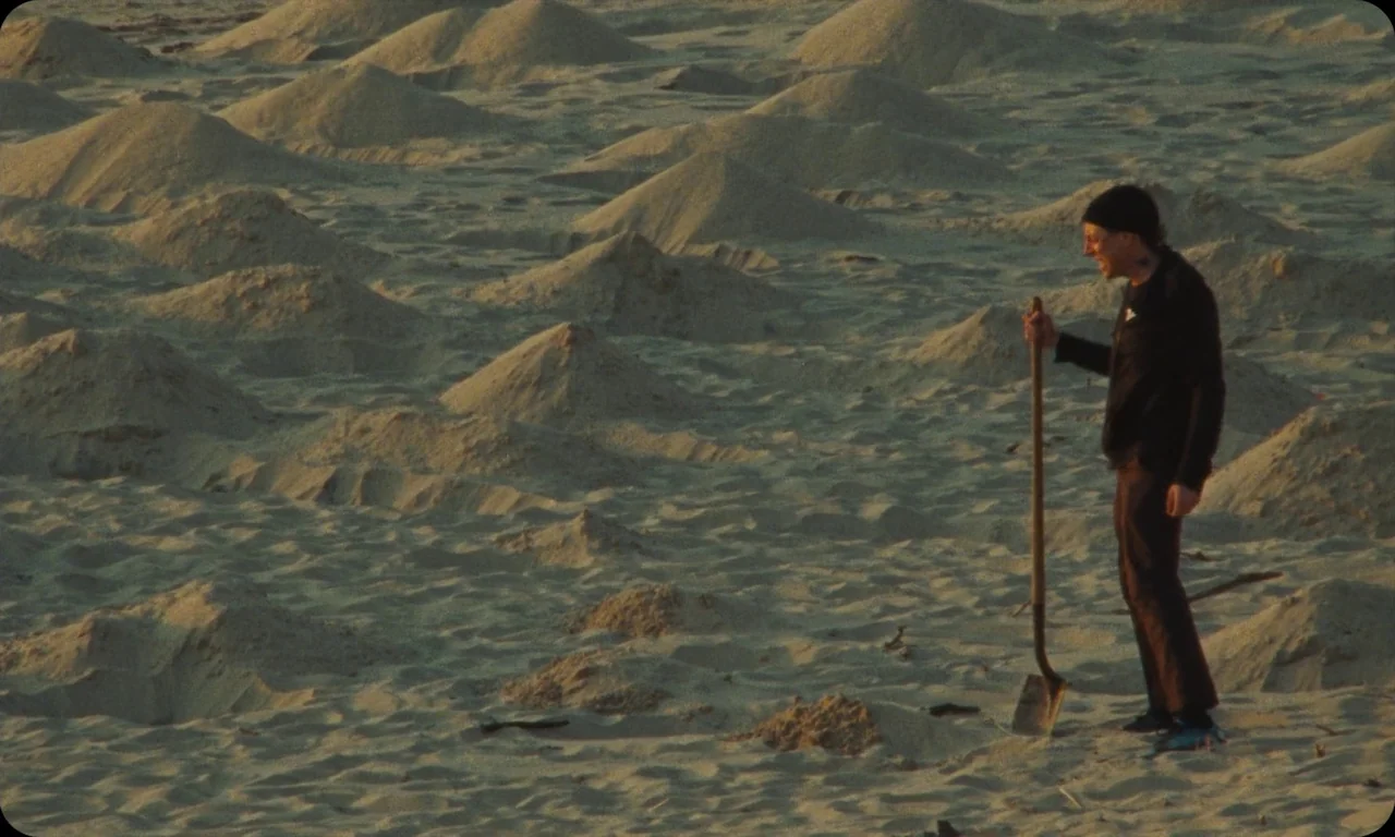 Vulcanizadora (2024) movie still: wide — A vast expanse of sand patterned with numerous small, uniform conical mounds. A man stands alone in the…