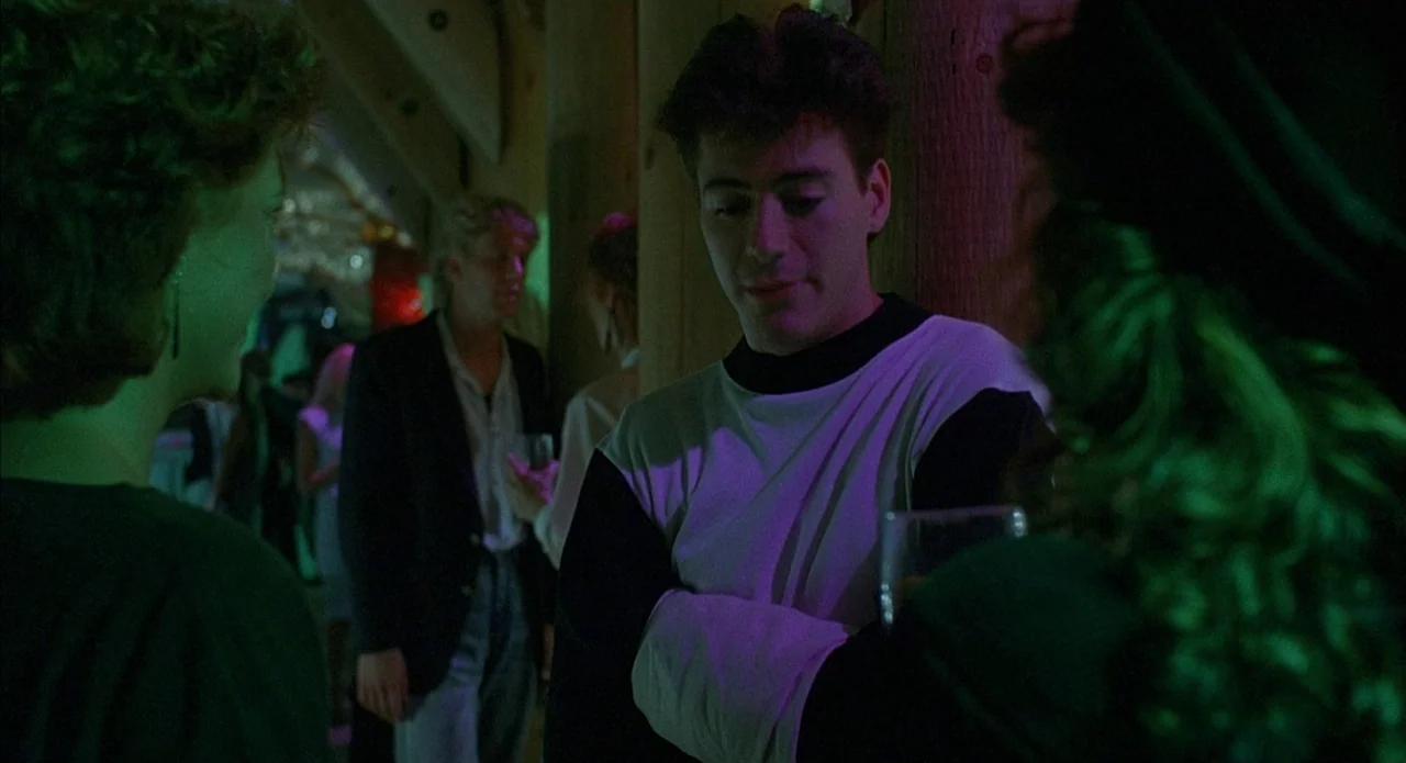 Less Than Zero (1987) movie still: medium — A dimly lit nightclub features wooden support beams and an crowded dance floor. The young man leans…