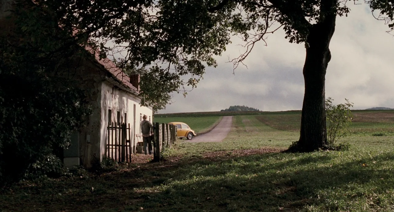 Revanche (2008) movie still: wide — A rural landscape features a weathered building and a single large tree. A person stands near a wooden fence…