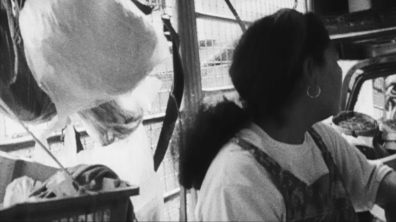 Mysterious Object at Noon (2000) movie still: medium — A woman stands in a busy market stall with hanging plastic bags and crates. She is looking to…