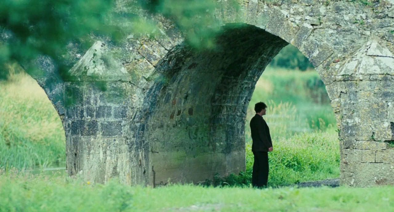 Jimmy’s Hall (2014) movie still: wide — An ancient moss-covered stone arch bridge spans a grassy meadow with green vegetation. A man stands in…