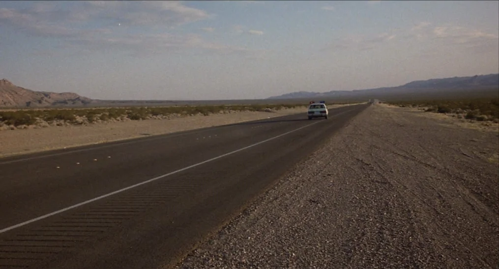 Salvador (1986) movie still: wide — An empty two-lane asphalt highway stretches through a vast, arid desert landscape. A lone white police cruiser…