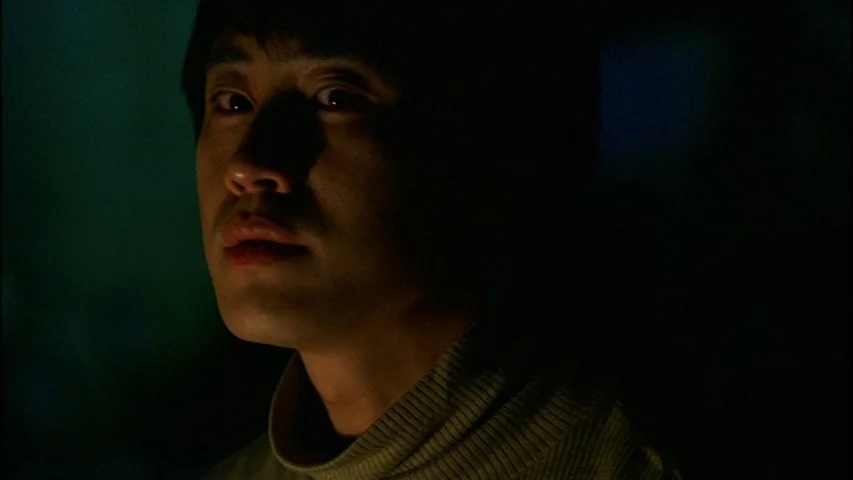 Save The Green Planet! (2003) movie still: closeup — A dark, dimly lit interior environment with minimal background detail visible. A young man is…