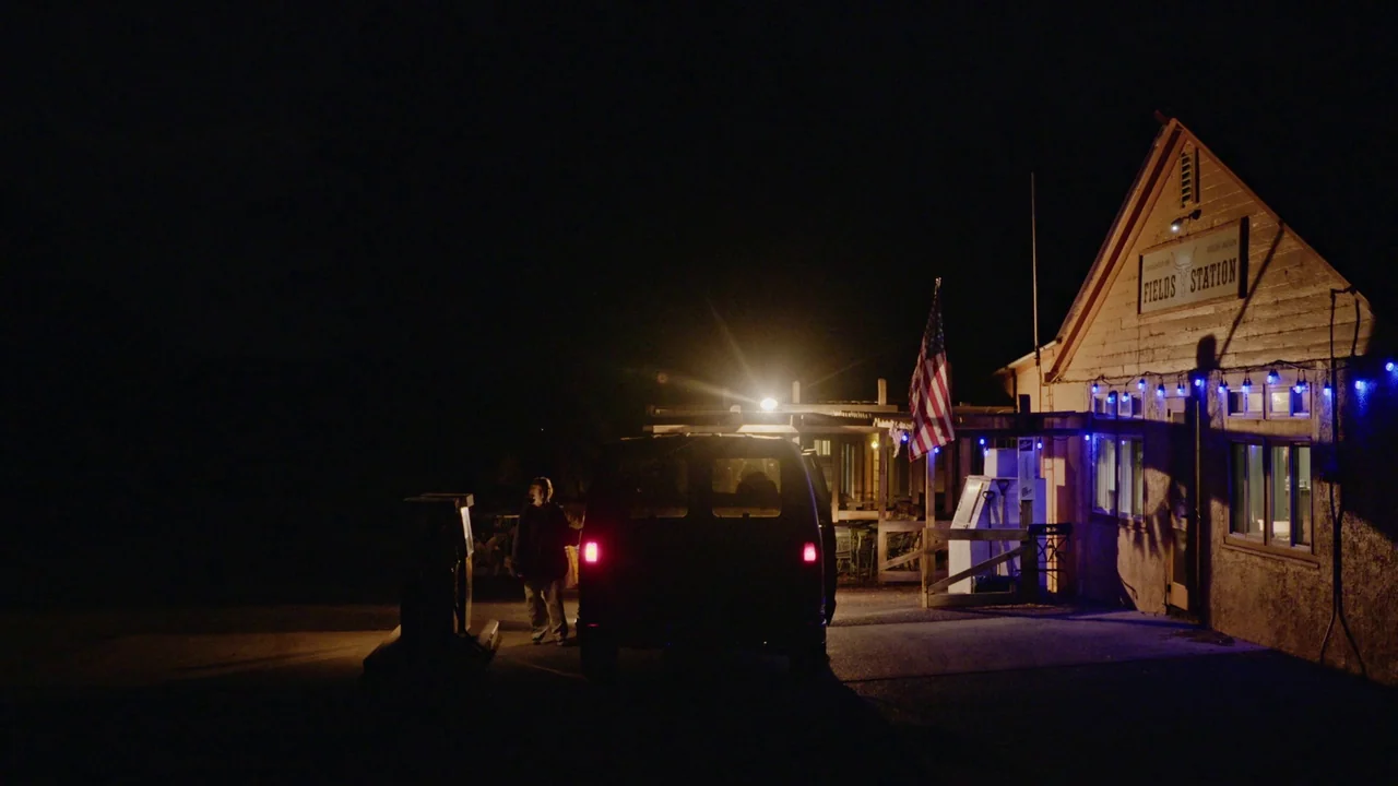 Gasoline Rainbow (2023) movie still: wide — A rural, wooden building labeled Fields Station stands in the dark of night. A lone man stands beside a…