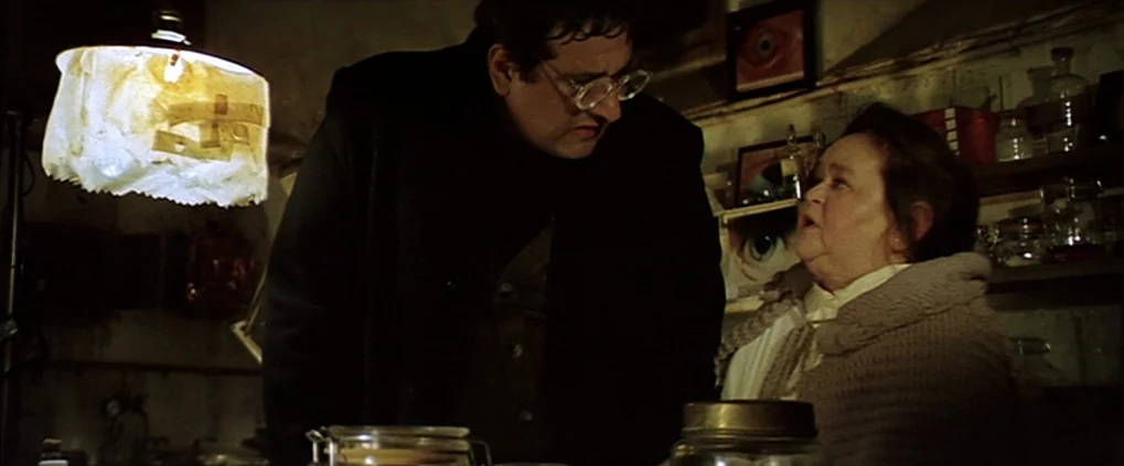 Anguish (1987) movie still: medium — A cluttered, dimly lit room contains numerous glass jars on shelves and artwork. A tall man in a dark coat…