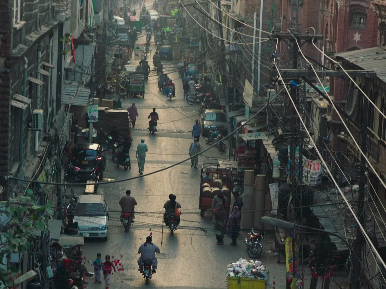 Joyland (2022) movie still: wide — A dense urban thoroughfare is crowded with motorcycles, pedestrians, and parked vehicles under tangled wires. The…