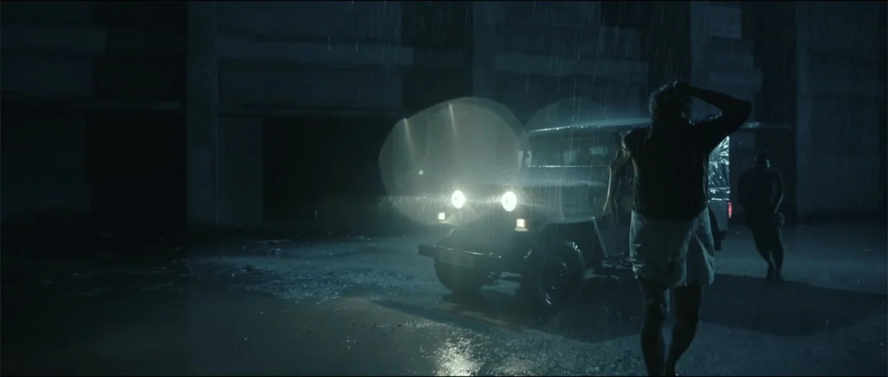 Garudan (2024) movie still: wide — A dark concrete lot is filled with heavy rain at night. A man runs toward the front of a waiting jeep with its…