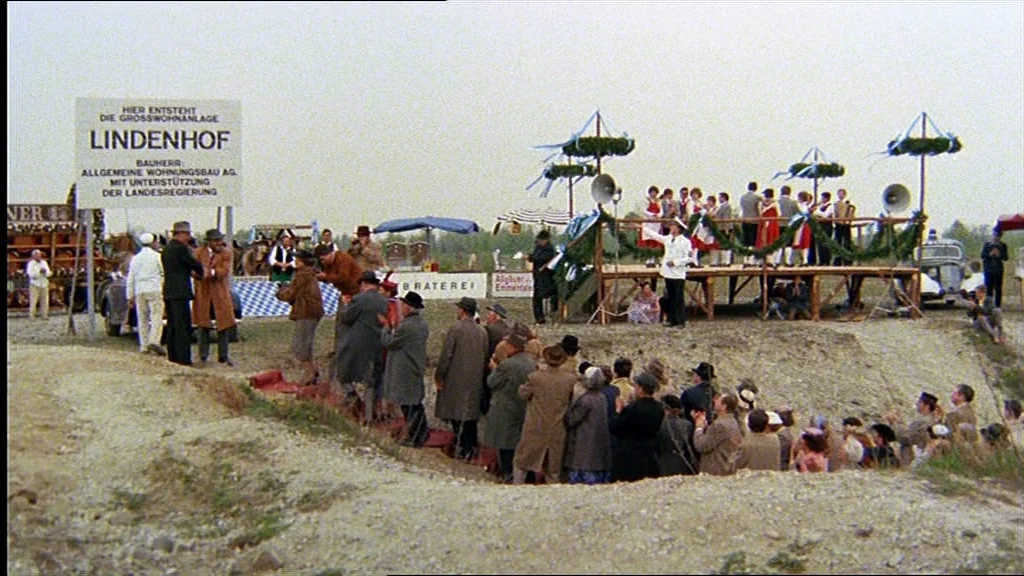 Lola (1981) movie still: wide — An outdoor construction site features a crowd of people gathered for an event. An elevated wooden stage with…