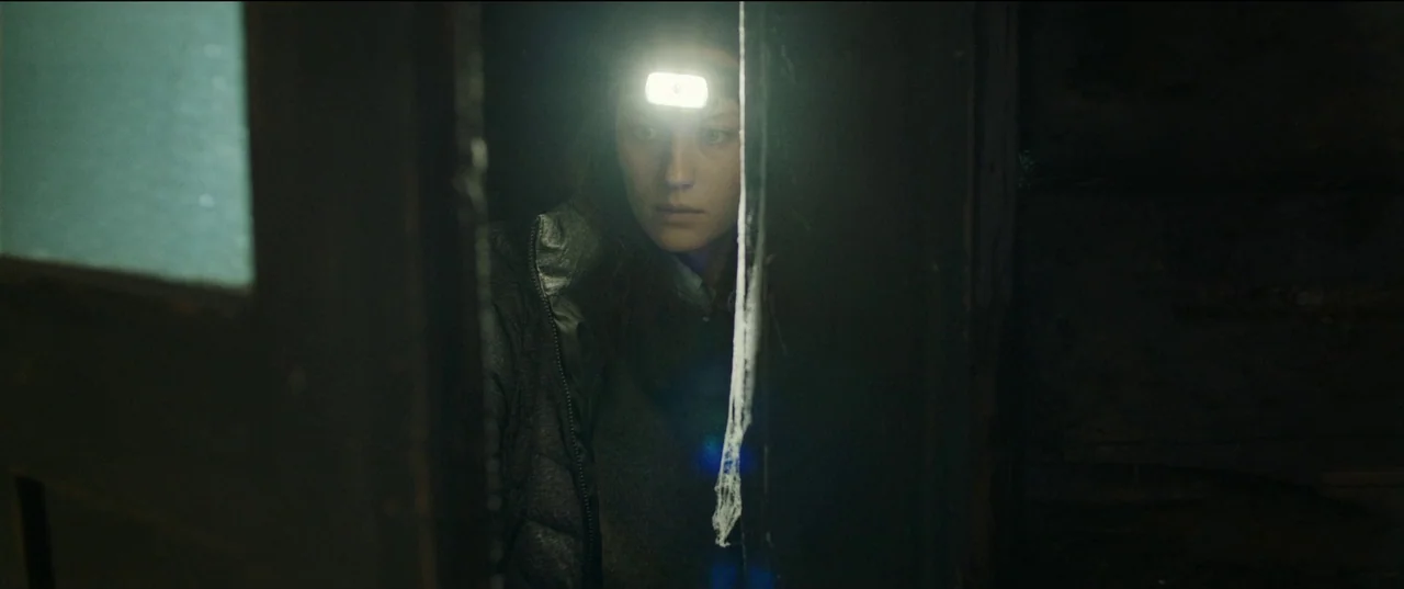 Nightsiren (2022) movie still: medium — A dark, narrow interior space with visible cobwebs hanging in the foreground. A woman wearing a headlamp…