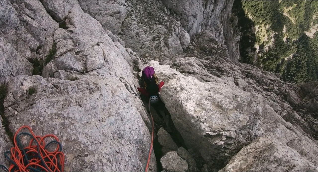 Crooked Trees Gon Give Me Wings (2022) movie still: wide — A vertical view looks down a steep, craggy mountain face made of grey rock. A lone…