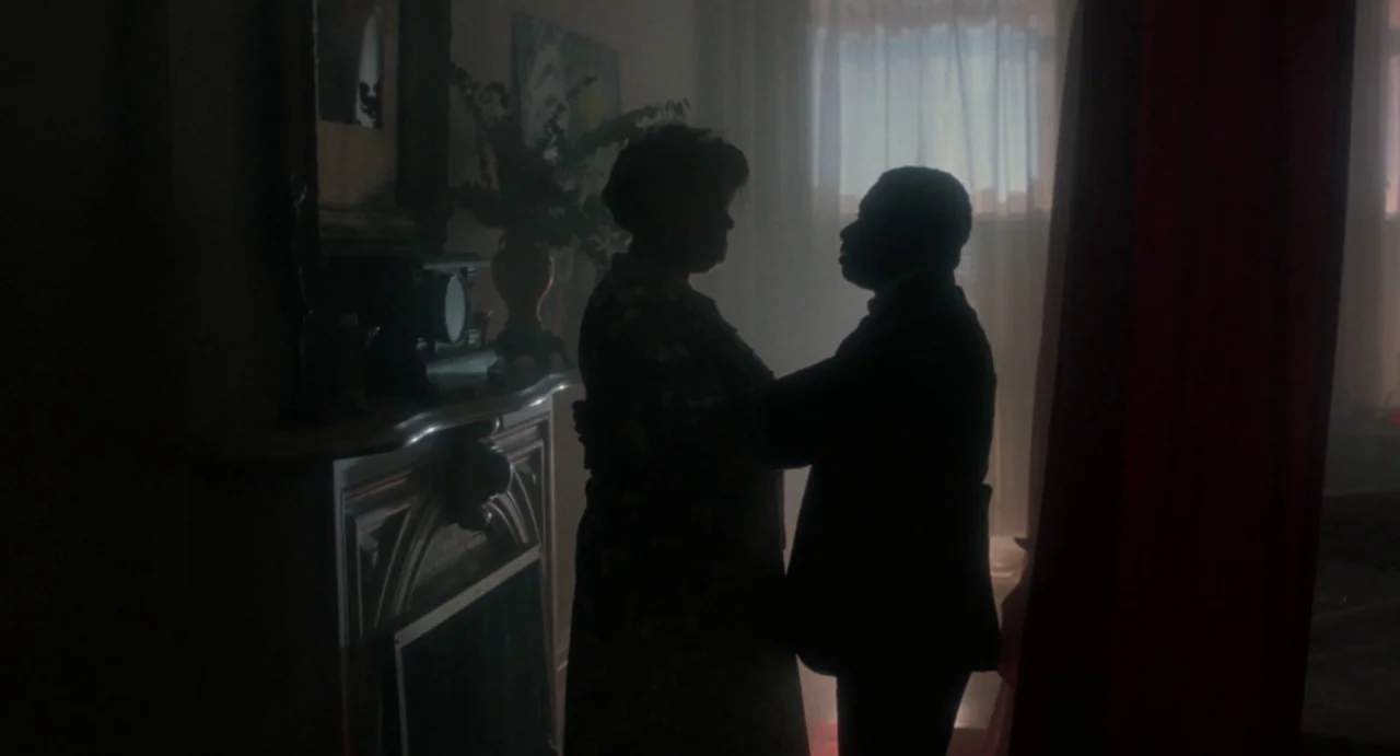 Def By Temptation (1990) movie still: medium — A dimly lit room with a fireplace and a window covered by sheer curtains. Two people embrace and sway…
