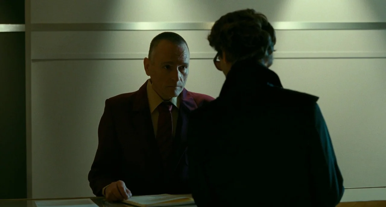 Saint Laurent (2014) movie still: medium — A sterile room features a reception counter against a pale, featureless wall. A man in a burgundy jacket…