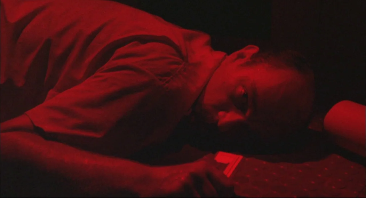 The Return of Godzilla (1984) movie still: closeup — A dimly lit room is filled with intense red light. A man lies flat on the floor in the…