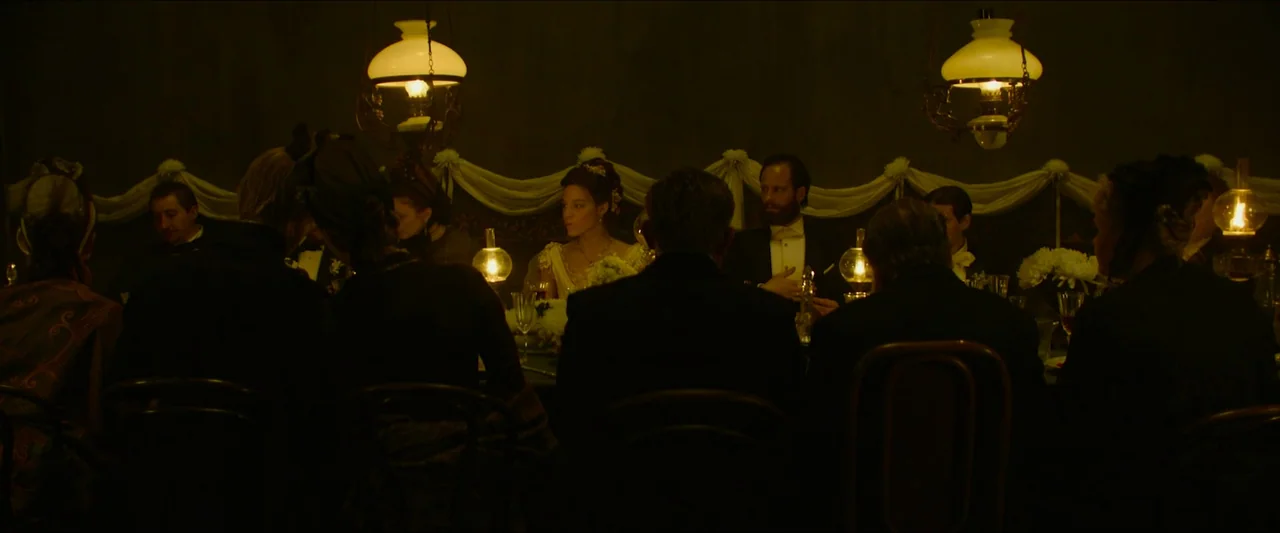 Tchaikovsky’s Wife (2022) movie still: wide — A dimly lit room contains a long dining table draped in white fabric cloth. Several people in formal…