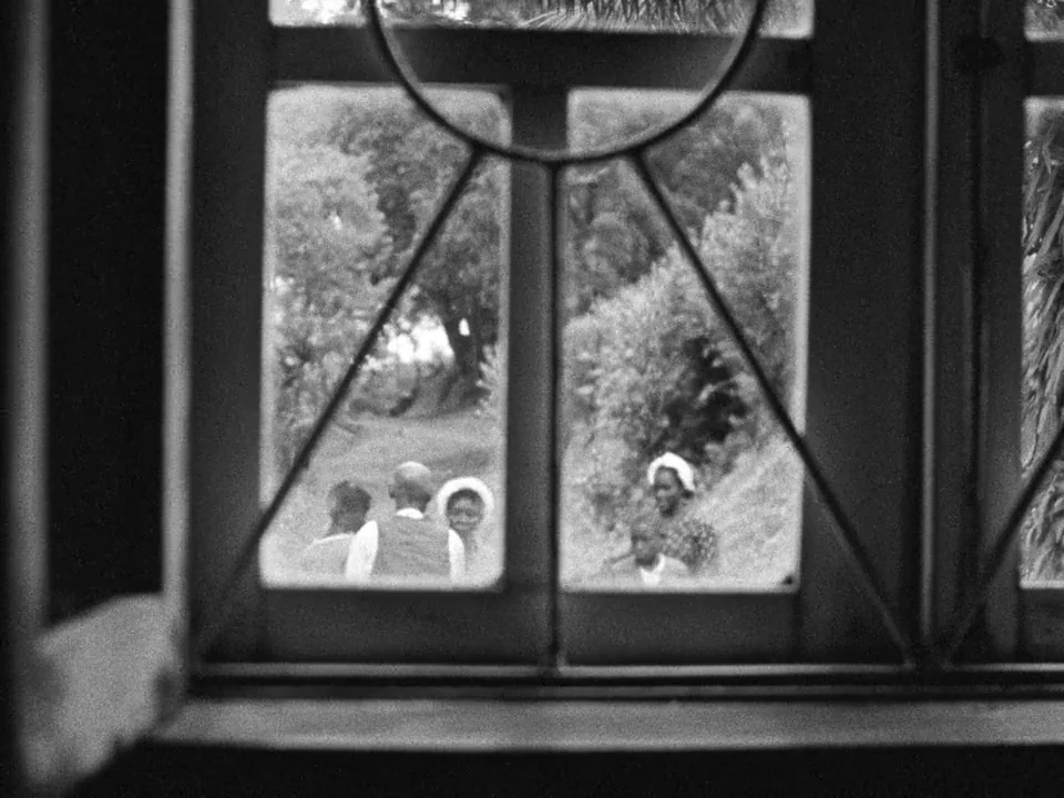 Tabu (2012) movie still: wide — A view through a paneled iron window looks out onto a leafy outdoor path. A small group of people walks along the…