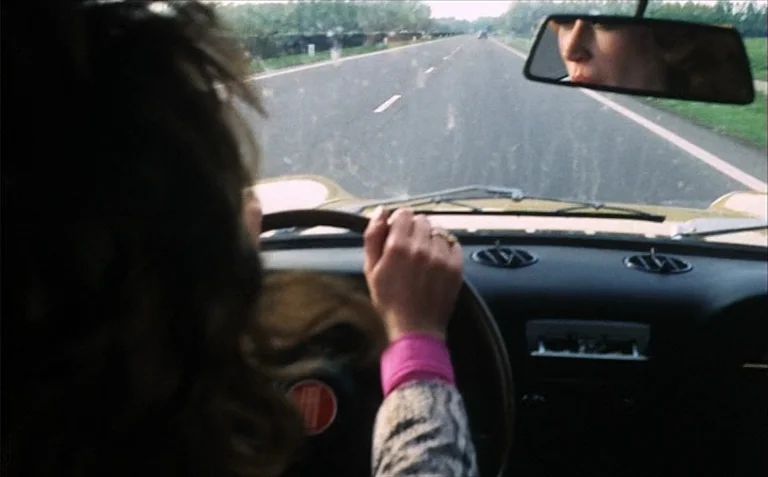 Business is Business (1971) movie still: medium — The interior of a car shows a person driving on a paved road. The rearview mirror reflects the…
