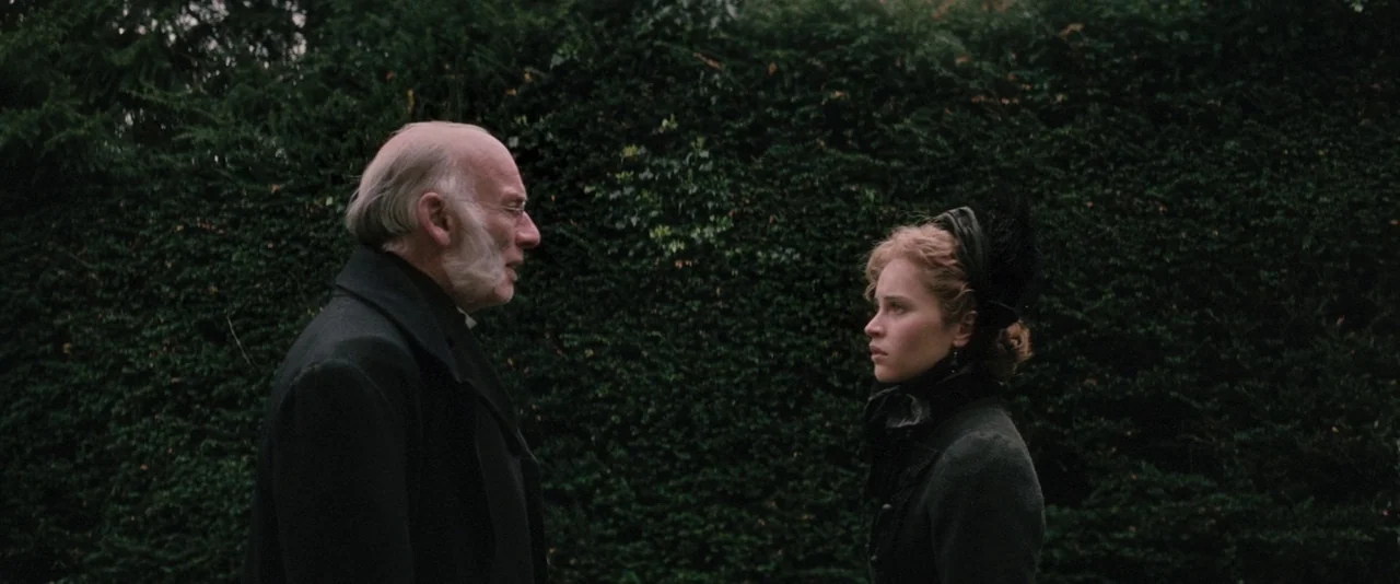 The Invisible Woman (2013) movie still: medium — Two individuals stand in profile before a dense, manicured green hedge. The older man speaks toward…