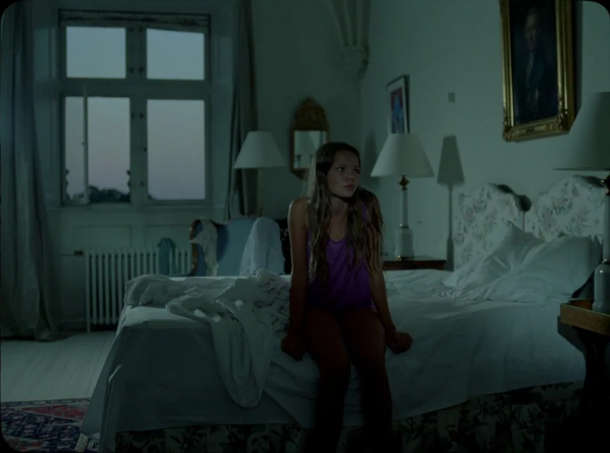 Jauja (2014) movie still: medium — A dimly lit room features a bed with white sheets and two bedside lamps. A teenage girl sits upright on the edge…
