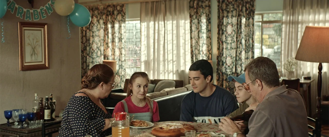 The Boy Who Killed My Parents (2021) movie still: medium — A family sits around a dining table in a house during the daytime. They are eating a meal…