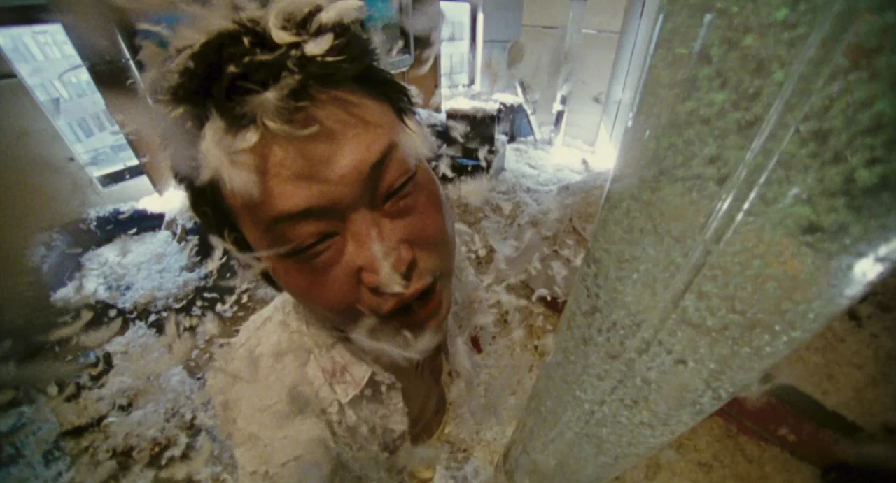 Ichi The Killer (2001) movie still: closeup — A messy, light-filled interior space is filled with thousands of floating white feathers. A man…