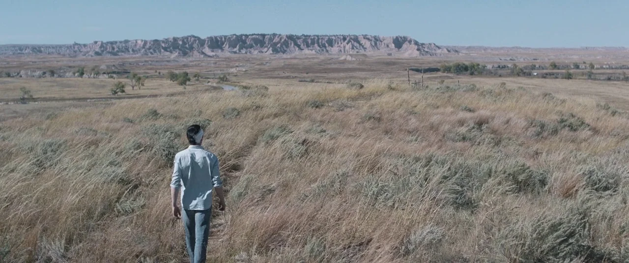 The Rider (2017) movie still: wide — A vast, dry grassland stretches toward a distant ridge under a pale blue sky. A man with a bandaged head walks…