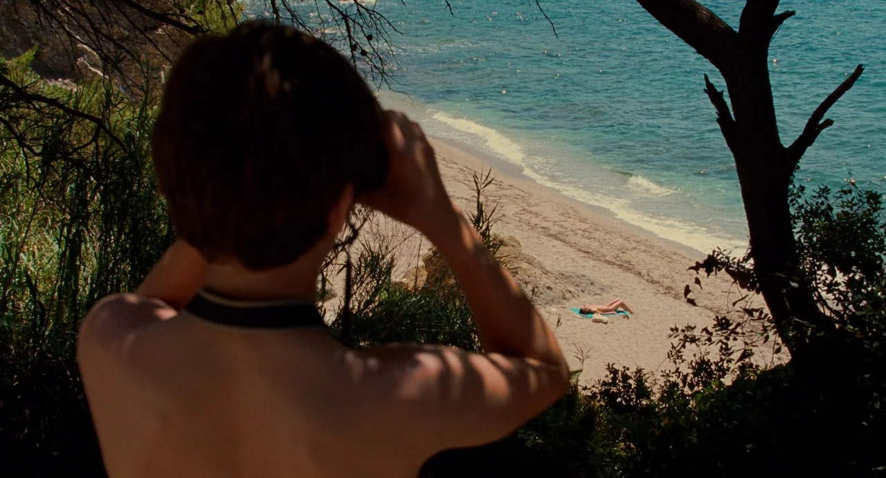 Young & Beautiful (2013) movie still: medium — A person viewed from behind peers through binoculars from a shaded, elevated coastal viewpoint. Far…