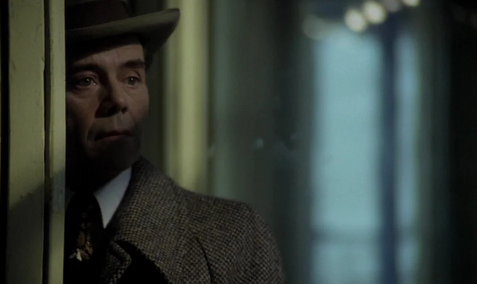 Despair (1978) movie still: closeup — A man looks through a doorway in a dimly lit, somber indoor space. He wears a tweed jacket and fedora while…