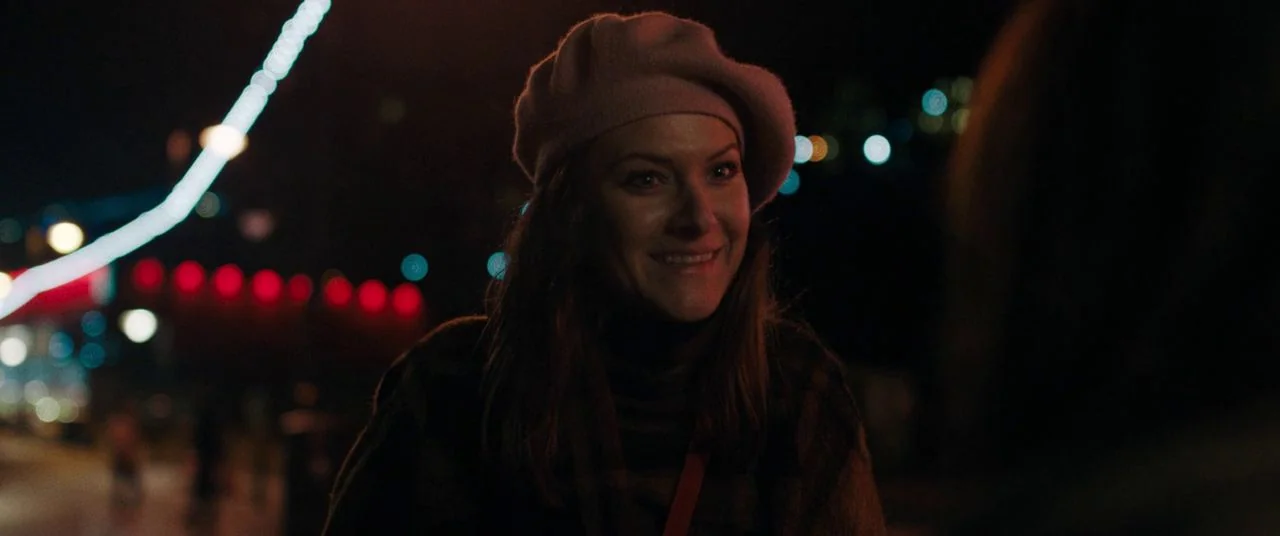 Saint Maud (2019) movie still: medium — A woman stands outdoors at night near an illuminated urban structure. She turns her face toward the camera…