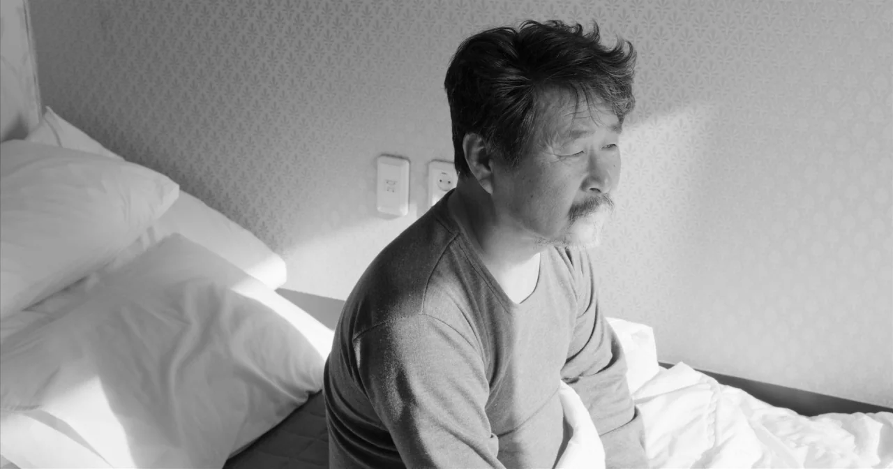 Hotel By The River movie still: medium — A white hotel room features patterned wallpaper and a neatly made bed. An elderly man in a gray shirt sits…