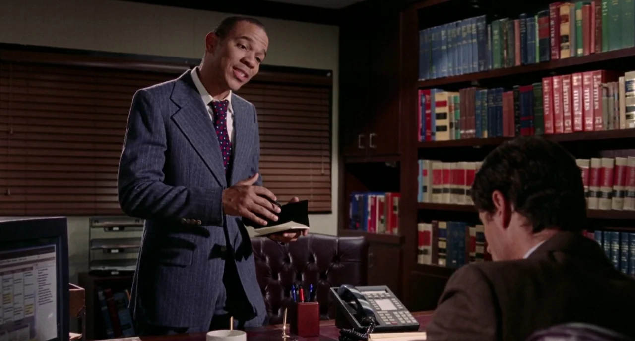 The Love Witch (2016) movie still: medium — A professional office with dark wood shelves filled with many books behind a desk. A man stands in front…