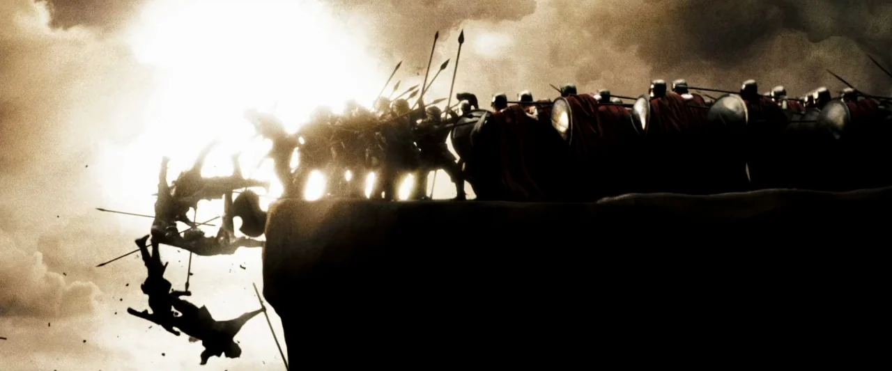 300 (2006) movie still: wide — A rocky cliff edge stands against a blinding white sky filled with clouds. A phalanx of soldiers in armor pushes…