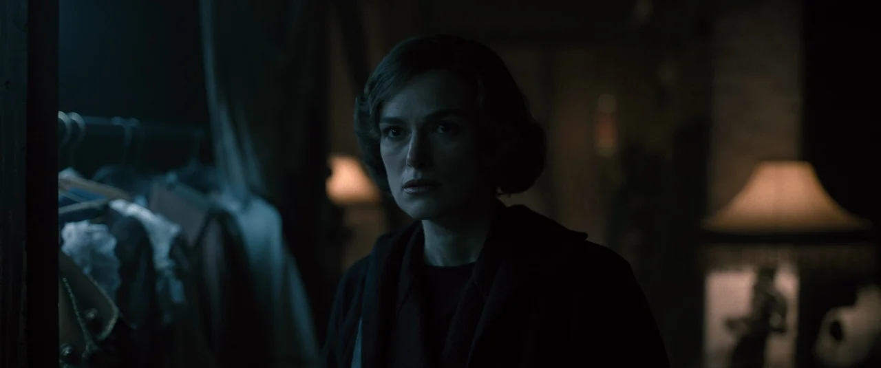 Boston Strangler (2023) movie still: medium — A dimly lit room with clothing racks and an out-of-focus table lamp. A woman with dark hair stands in…