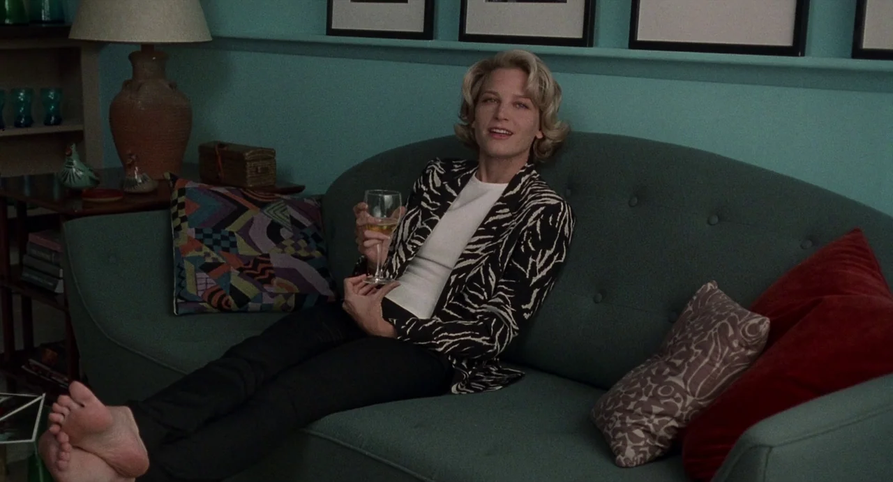 Touch (1997) movie still: medium — A teal-walled room contains a dark green sofa adorned with colorful throw pillows. A blonde woman sits…