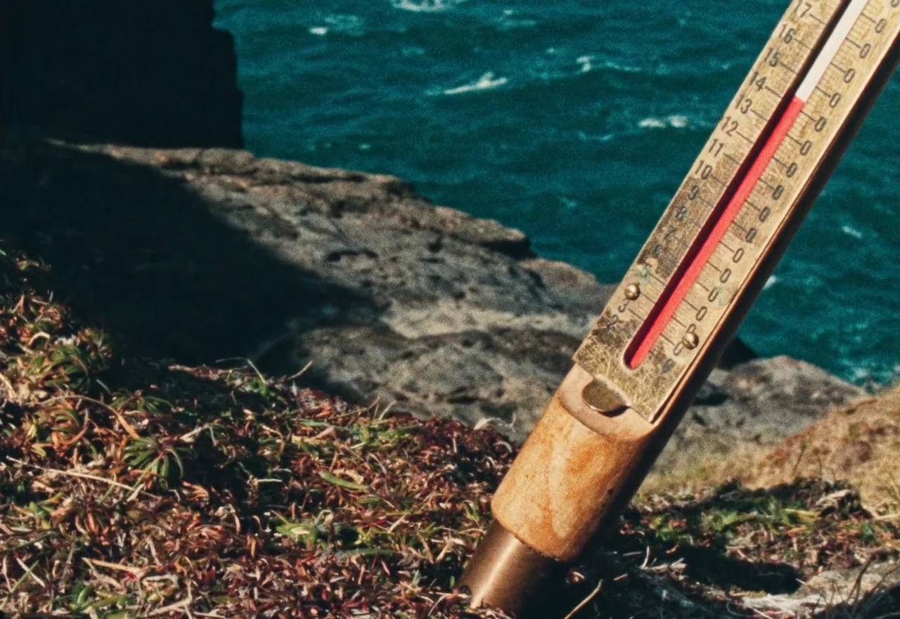 Enys Men (2022) movie still: closeup — A brass-framed thermometer is anchored in the dirt of a rocky coastal cliff. The deep blue sea is visible in…