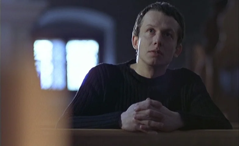 Blind Chance (1987) movie still: medium — A dark, dimly lit interior space with rows of wooden seating pews. A man sits at a pew, hands clasped,…
