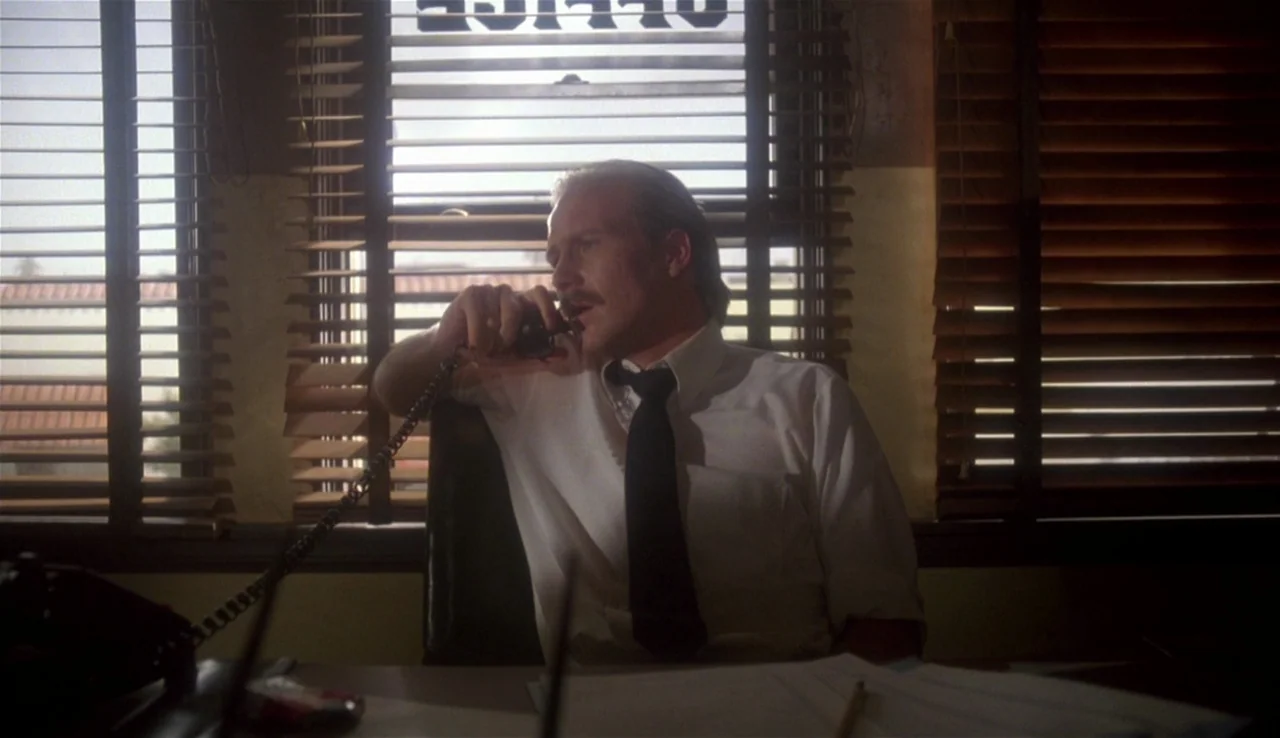 Body Heat (1981) movie still: medium — A man sits behind a desk in front of large windows with brown venetian blinds. The word OFFICE is printed…